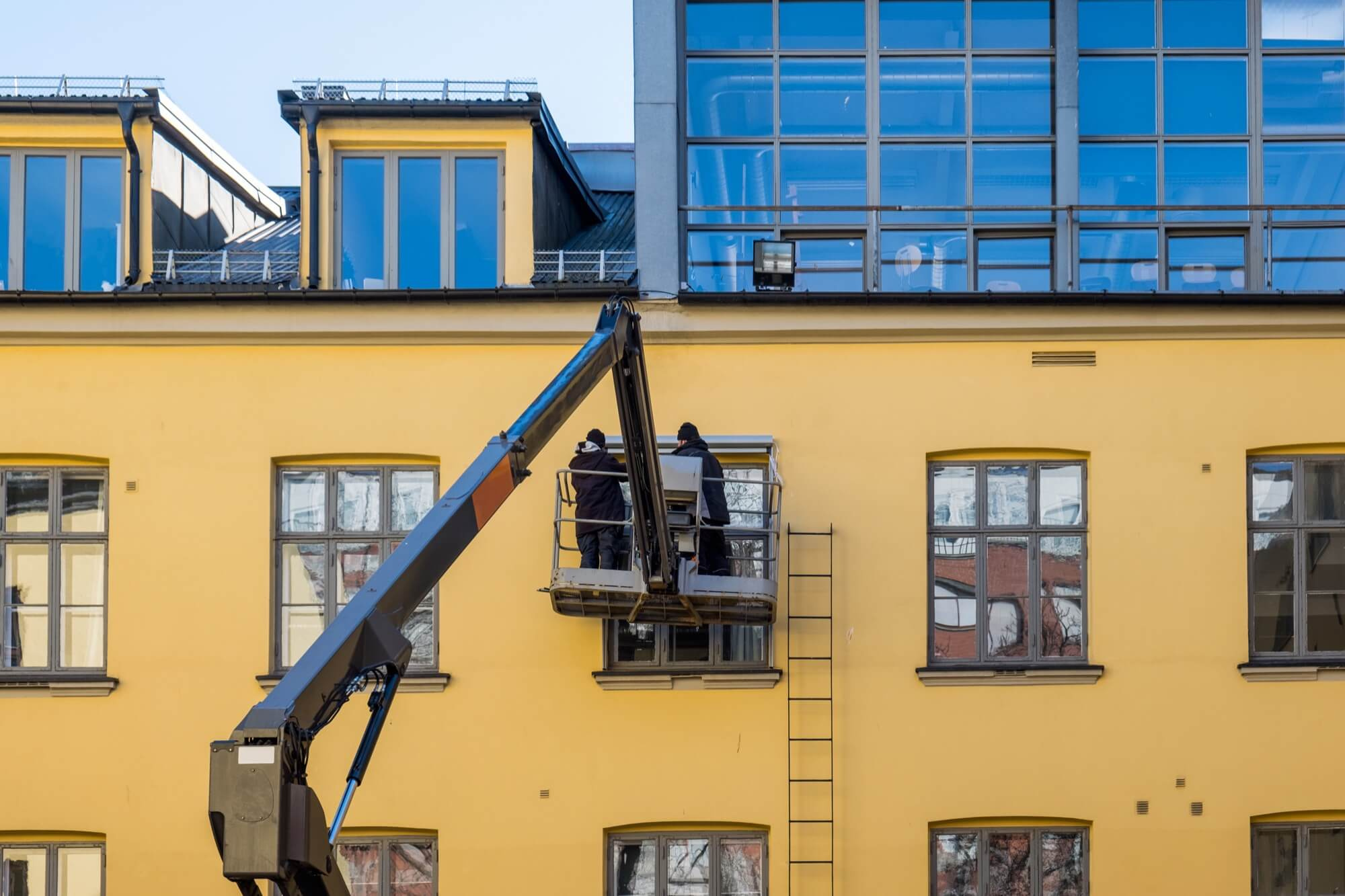 Två hantverkare utför fasadrenovering med lift på en gul byggnad med flera fönster.