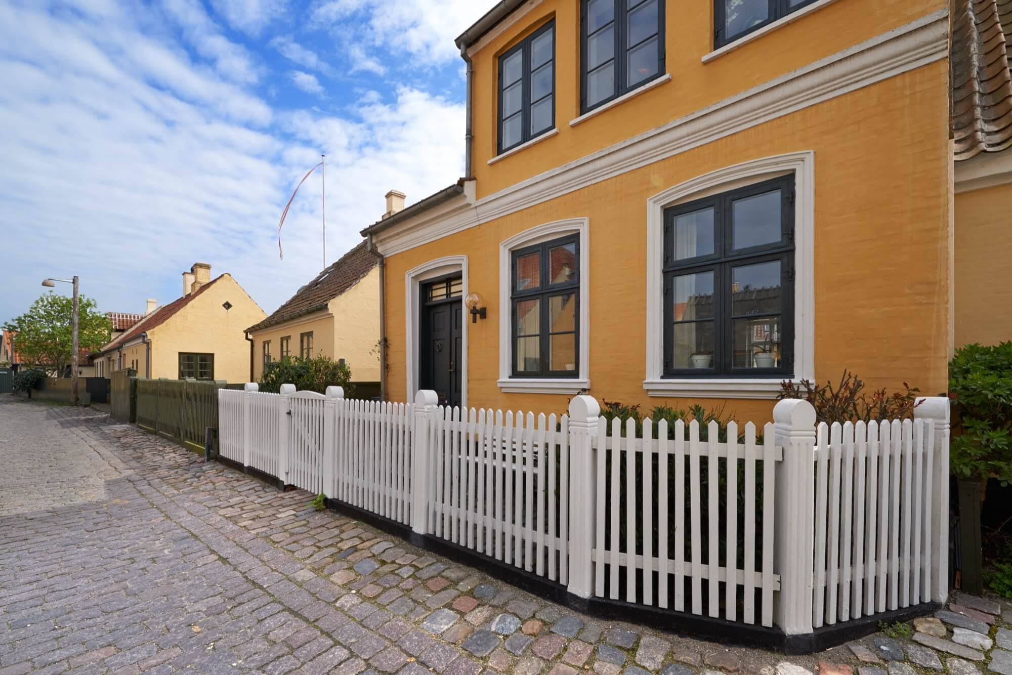Nyrenoverad gul fasad på ett bostadshus med vitt staket längs en kullerstensgata.
